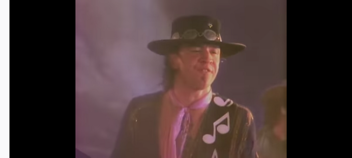 Stevie Ray Vaughan and DOUBLE TROUBLE video for Couldn't Stand the Weather.  Stevie Ray, white man with shoulder length dark hair, is wearing his signature flat black cowboy hat, black jacket, black strap of guitar with large white notes over his shoulder, and pink hankerchief tied around his neck.