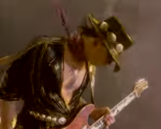 Stevie Ray Vaughan and DOUBLE TROUBLE video for Couldn't Stand the Weather.  Stevie Ray, white man with shoulder length dark hair, is wearing his signature flat black cowboy hat, black jacket, black strap of guitar with large white notes over his shoulder, and pink hankerchief tied around his neck.  He is leaning to his left into the wind and pouring rain. He is looking down with his hat pulled down to cover his face, holding his guitar up with his left hand, and his hankerchief is blowing behind him.