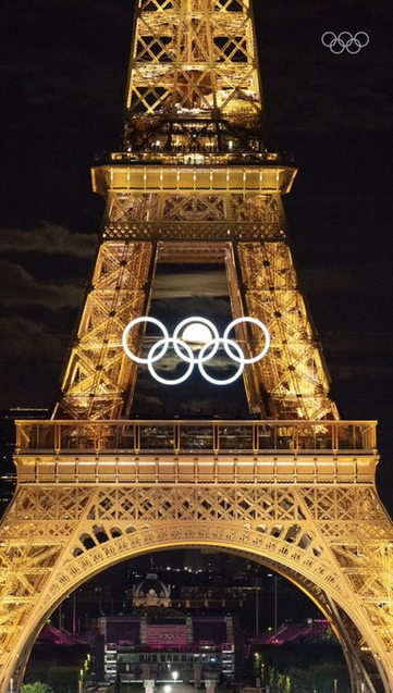 The images depict the Eiffel Tower at night, illuminated in golden light. Prominently displayed in the center of the tower are the Olympic rings, glowing brightly. In the background of the rings, the moon is perfectly positioned, creating the illusion that it is part of the rings. Zoom in to see how perfect the moon fits in the middle ring. It's almost like it's highlighted, or doesn't want to be left out or ignored. 