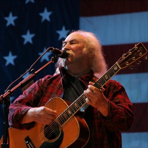 David Crosby singing with a guitar in front of an American flag.