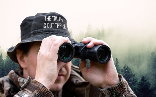 Man dressed in camo using binoculars in foggy forest wearing a hat reading 